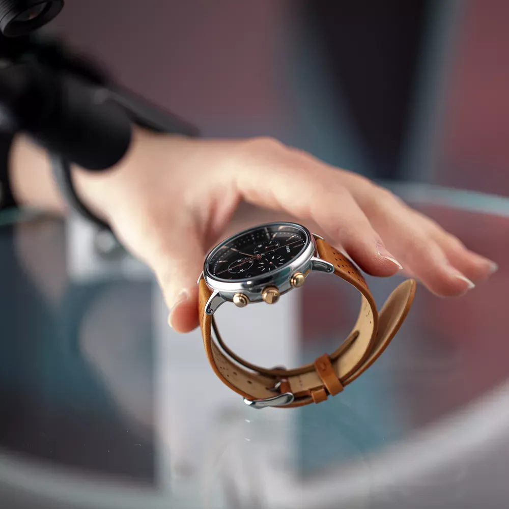 Mano colocando un reloj de hombre con correa de cuero marrón sobre una superficie de cristal